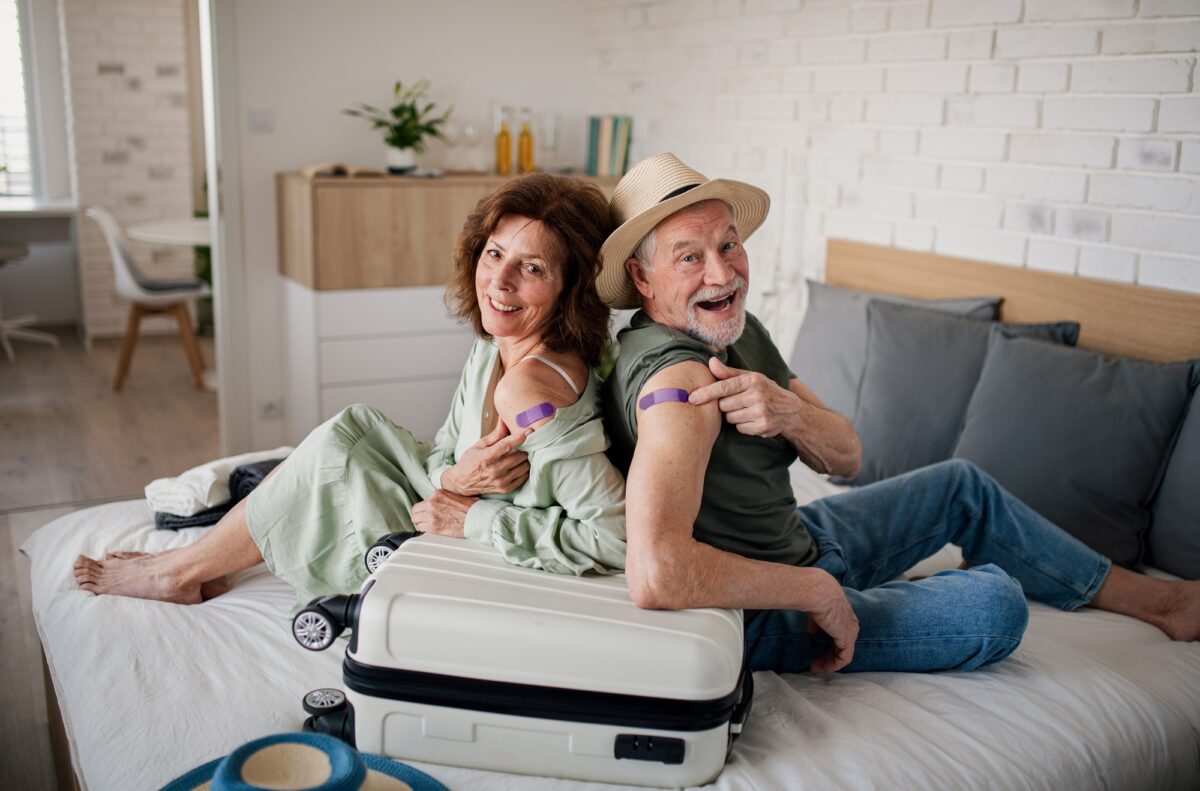 Senior couple sitting on bed indoors in hotel room, coronavirus vaccination and holiday concept.