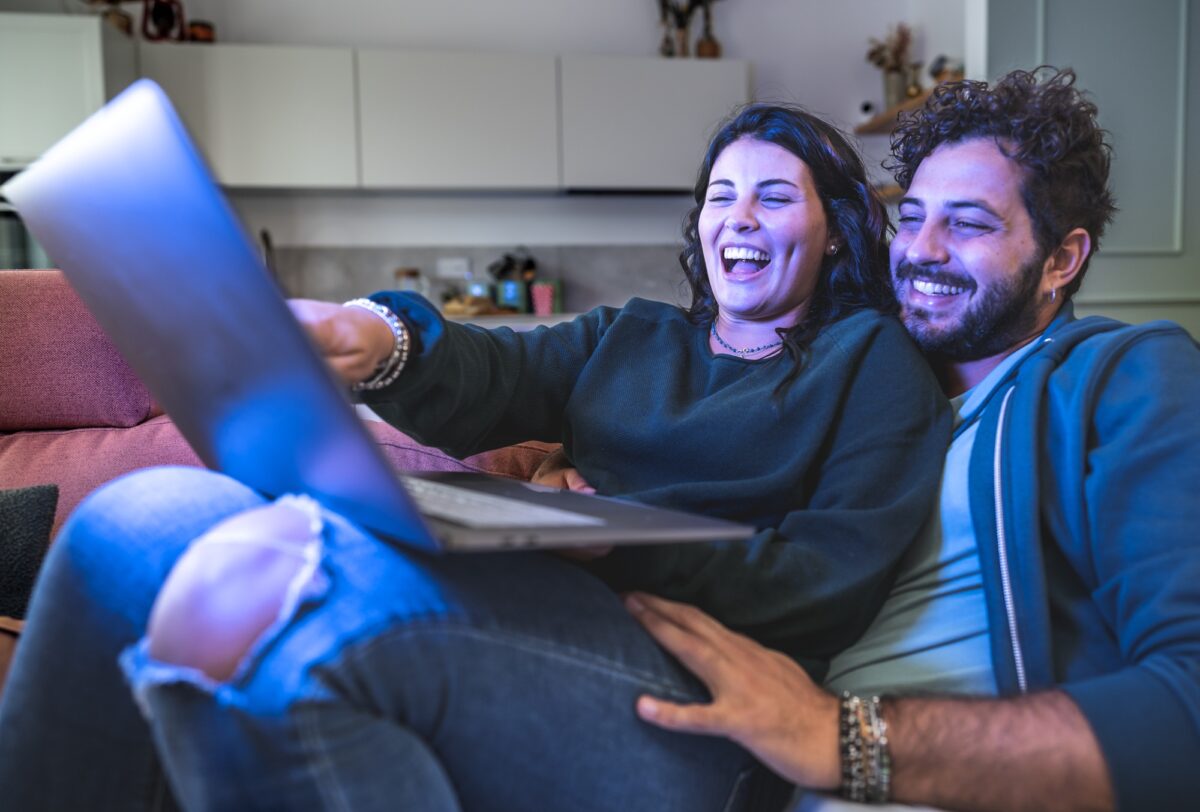 A young couple cuddles on a cozy couch as they search for budget-friendly travel deals on a laptop.